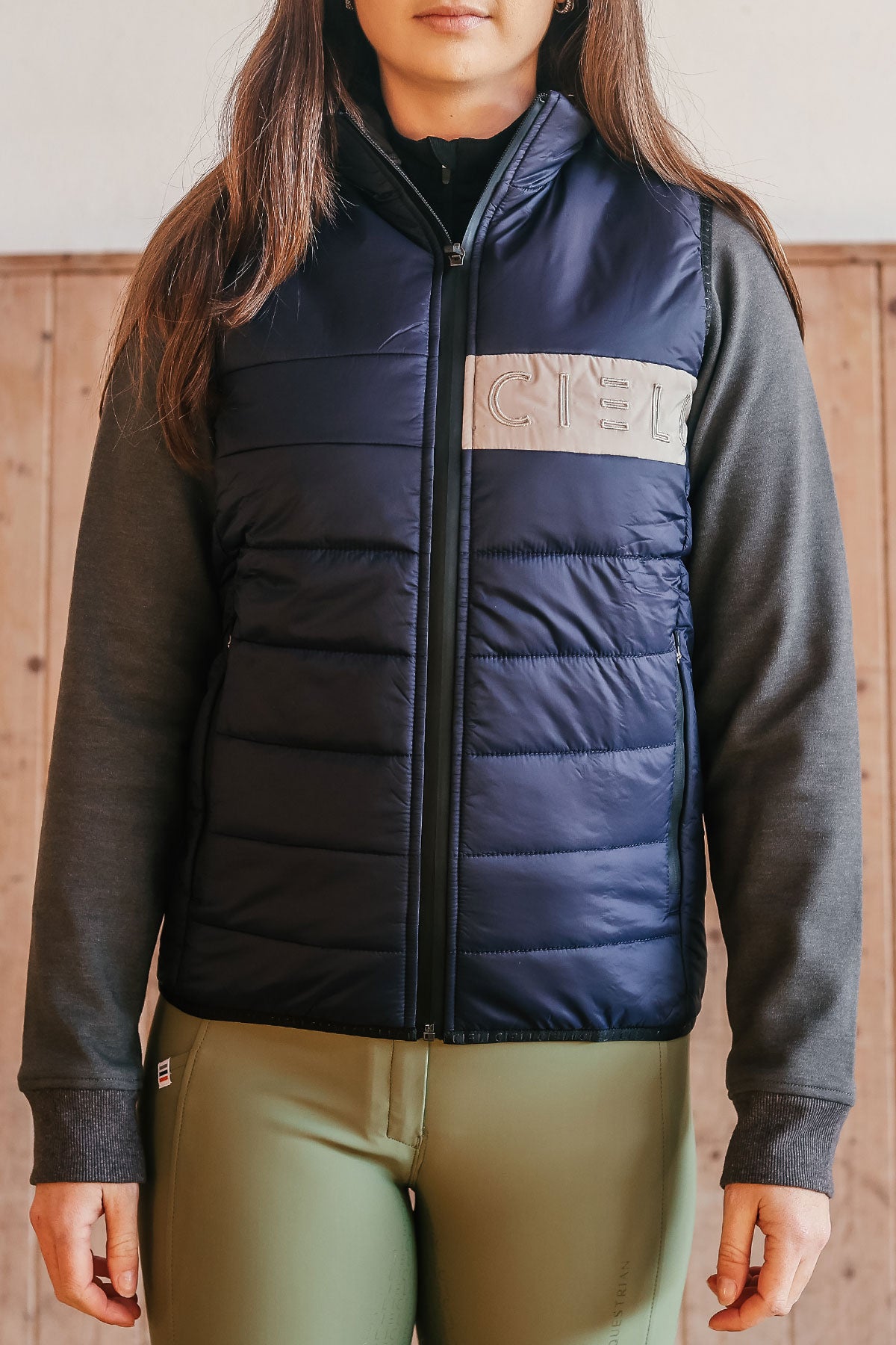A person wearing a navy Carter Body Warmer vest with a cream Cieli logo patch on the chest, paired with a grey long-sleeved hoodie.