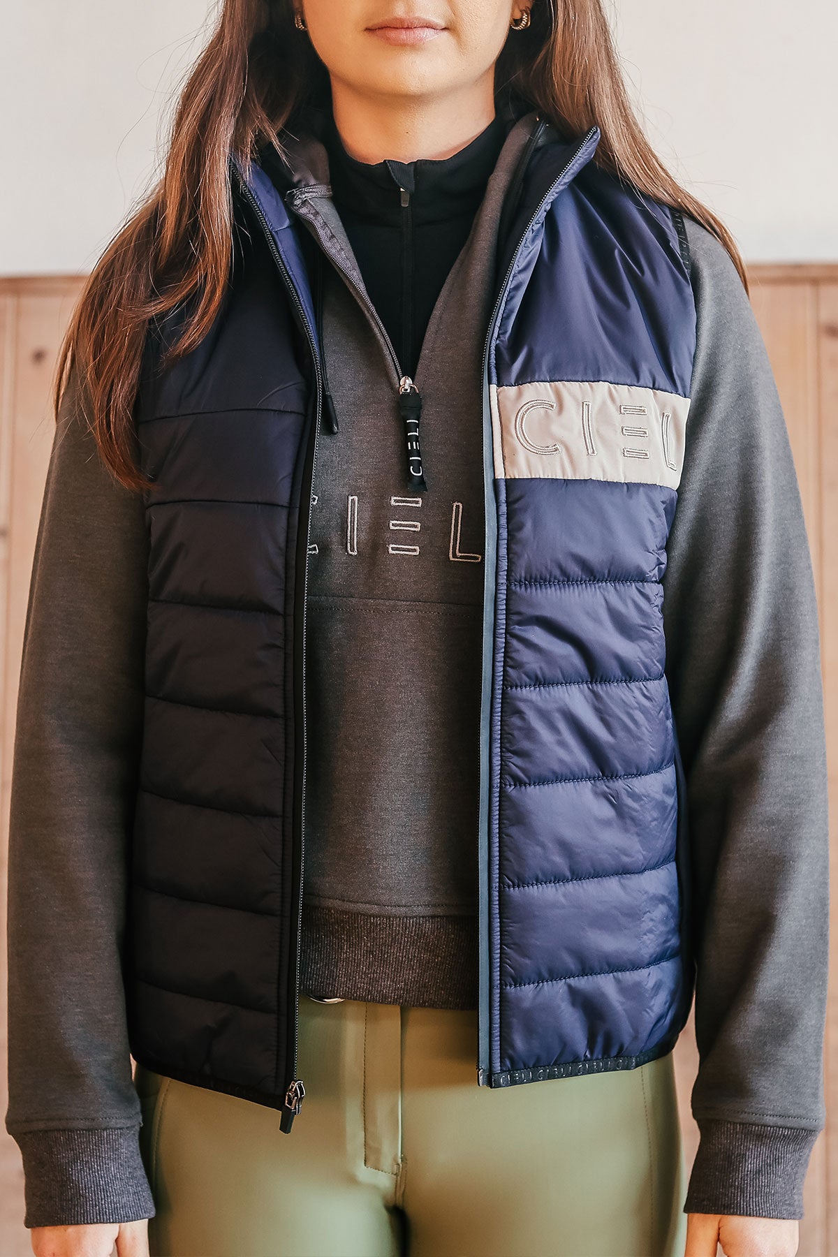 A person wearing a navy Carter Body Warmer vest with a cream Cieli logo patch on the chest, paired with a grey long-sleeved hoodie.