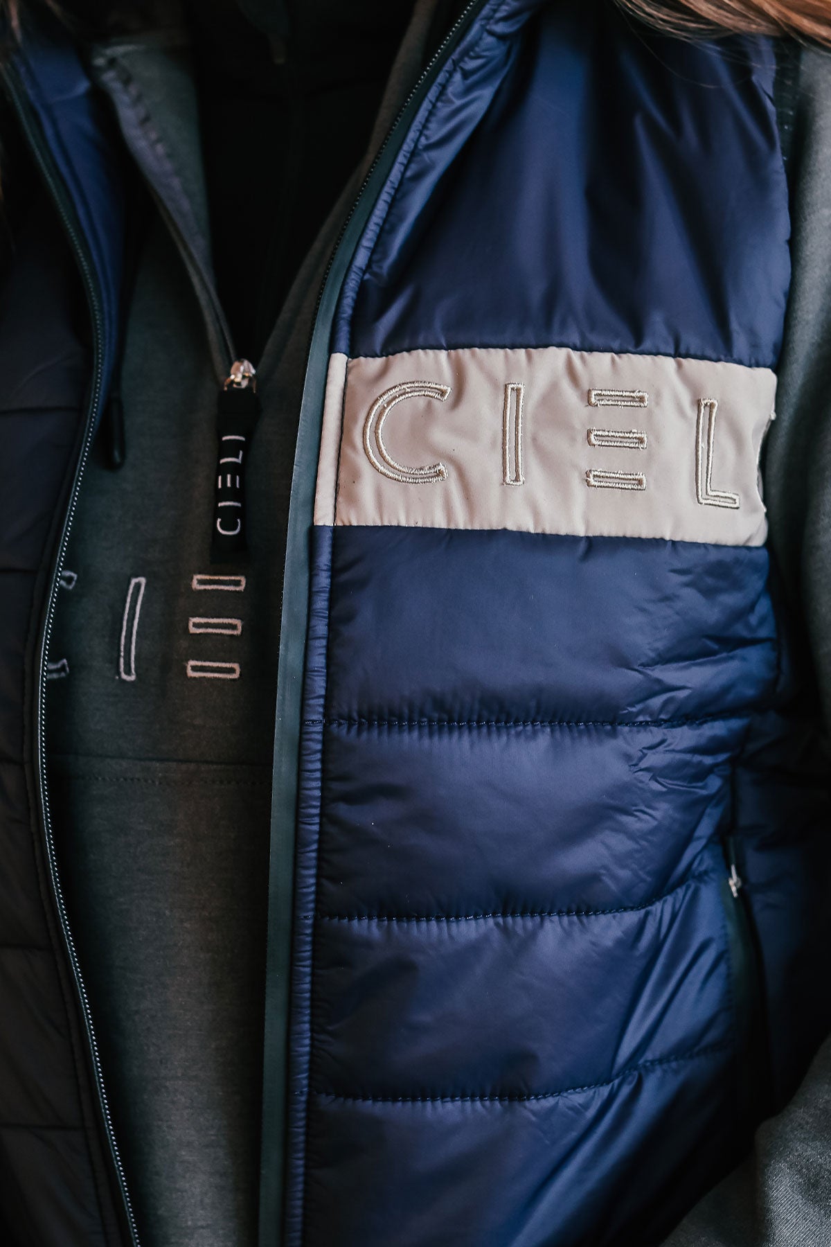 Close up of a person wearing a navy Carter Body Warmer vest with a cream Cieli logo patch on the chest, paired with a grey long-sleeved hoodie.