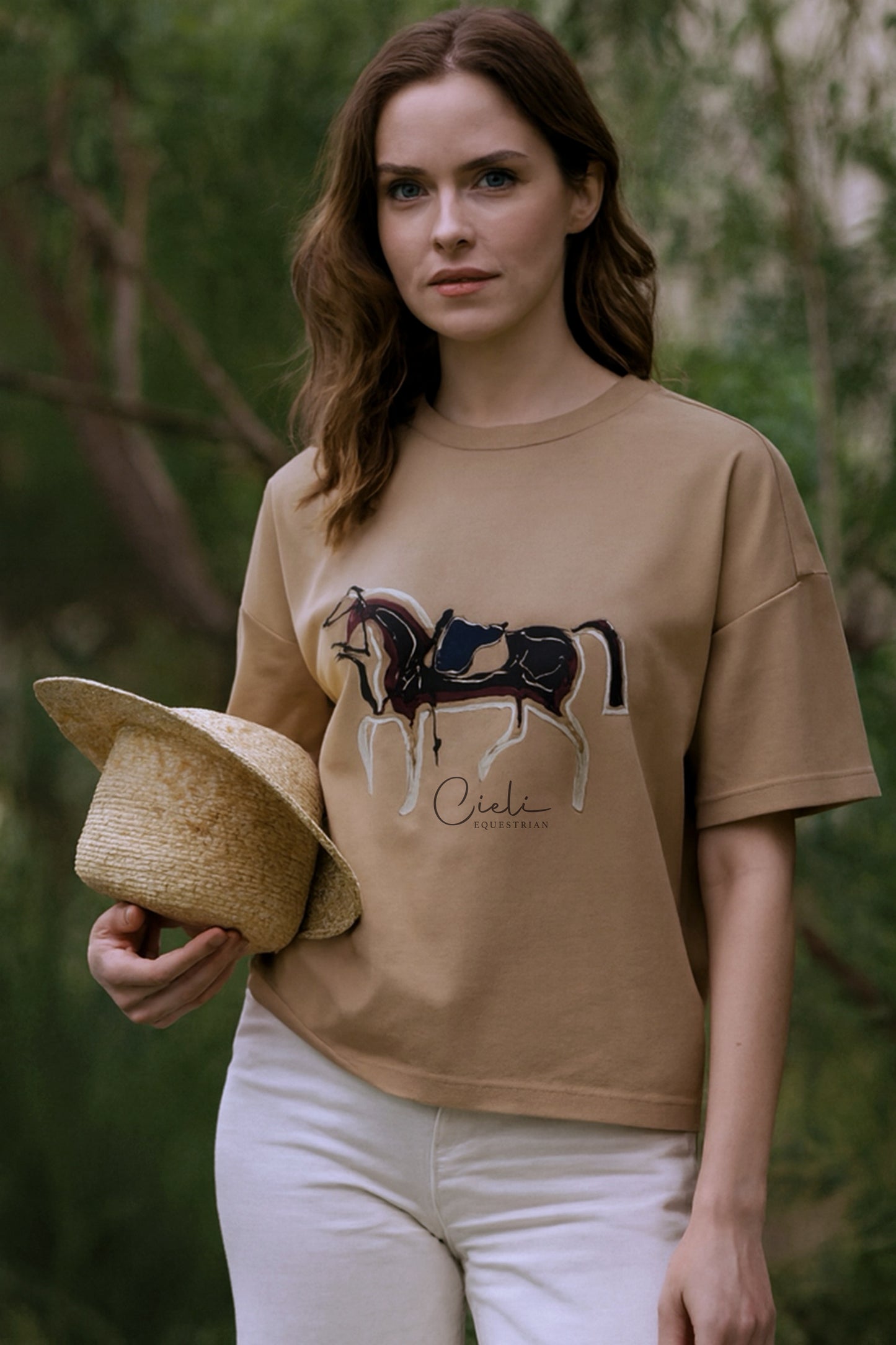 Woman wearing a beige t-shirt with a horse graphic and text, holding a hat in a forest setting.