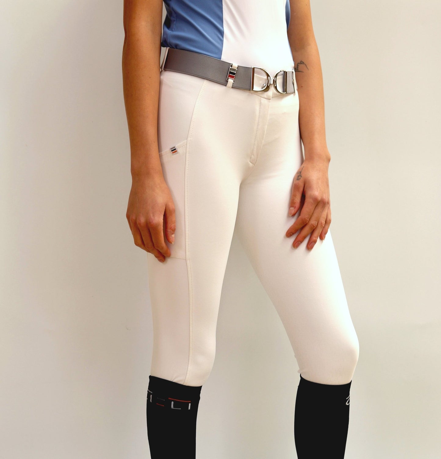 A woman modeling Berlin Performance Breeches by Cieli Equestrian. The breeches are white with orange accents, featuring a cell phone pocket with a logo label, belt loop with a pendant, and a subtle silicone print on the left leg.