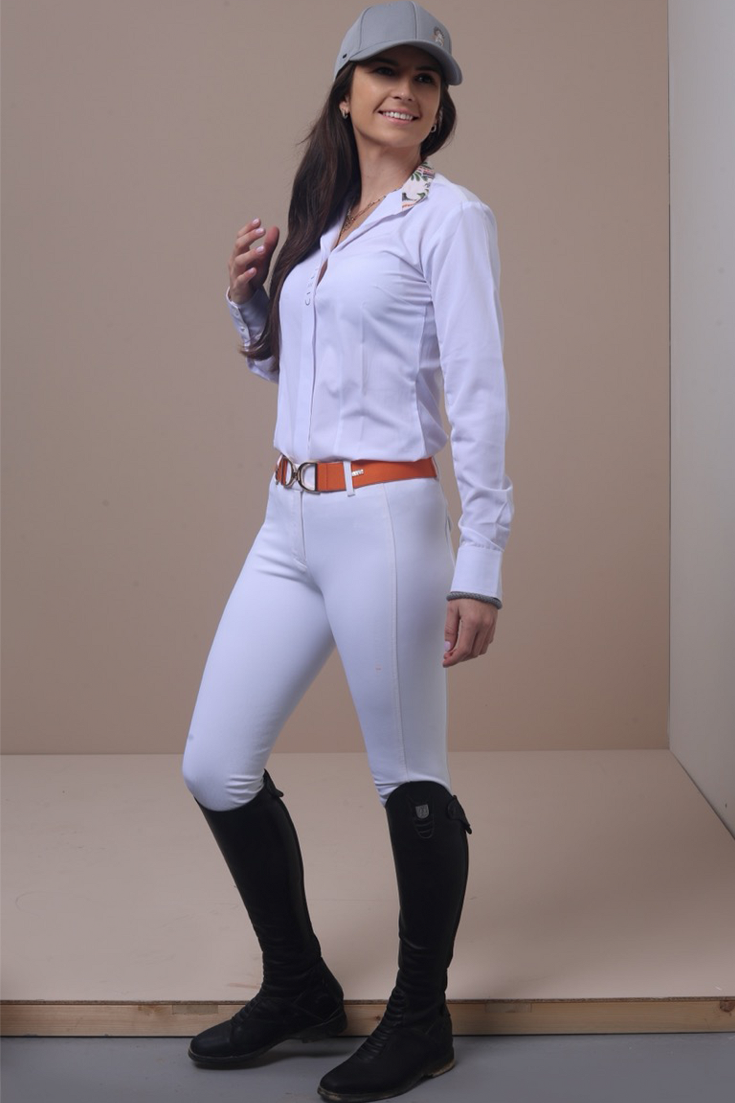 Woman in equestrian attire with white shirt, pants, and black boots against a plain background