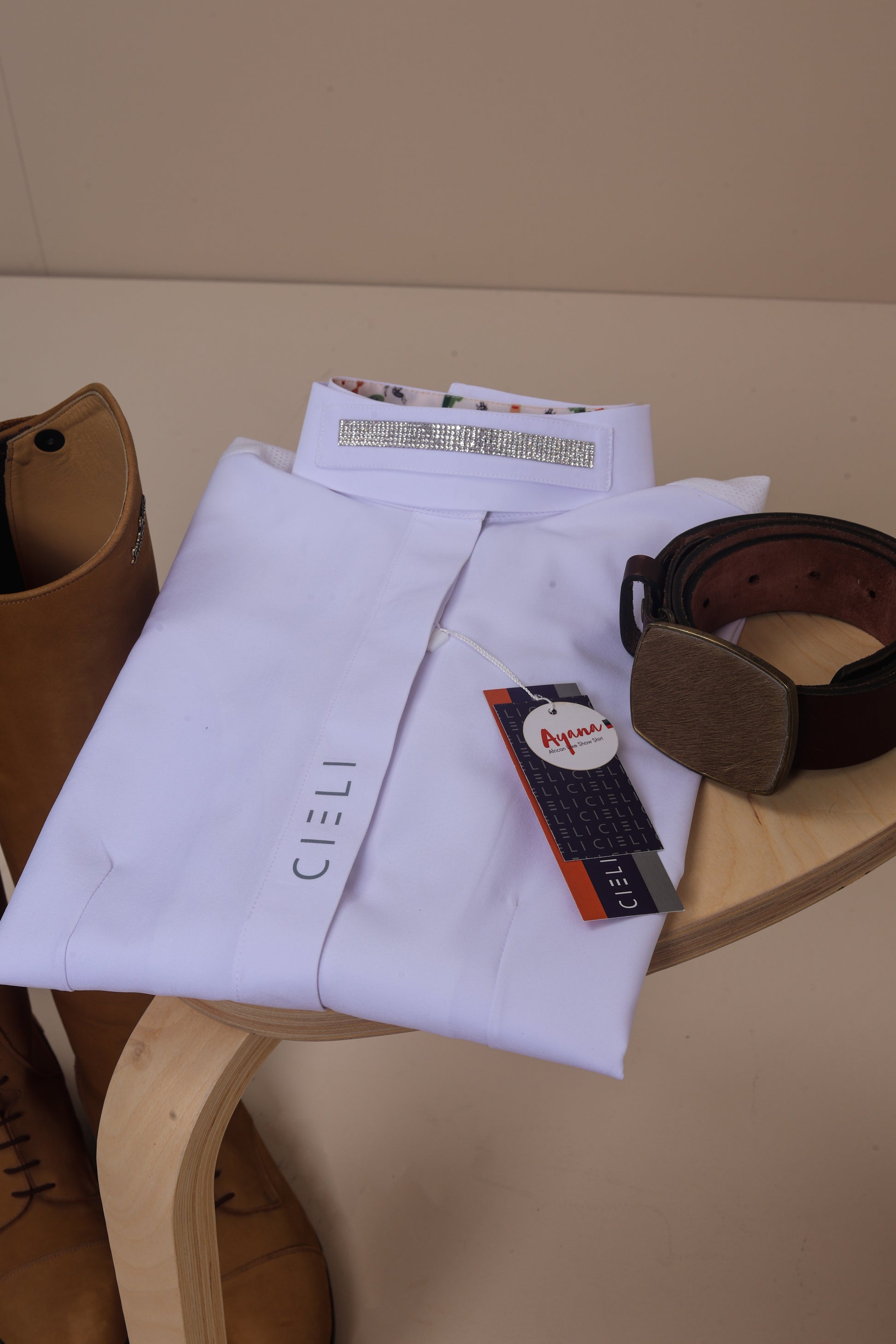 White shirt with a tag on a wooden stool, with a brown belt and riding boots in the background.