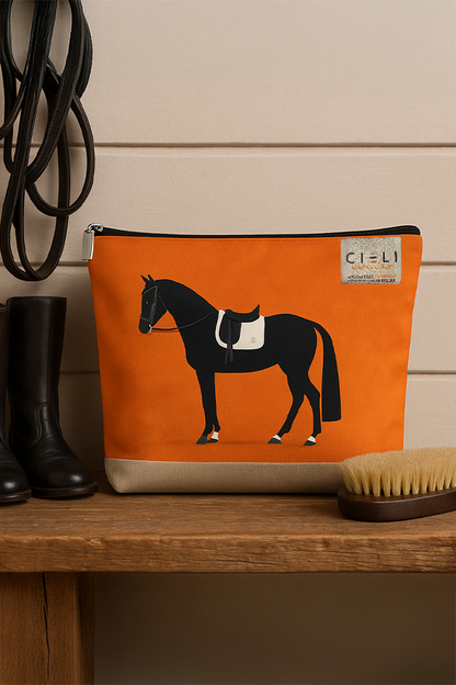 Orange pouch with horse design on a wooden surface next to black boots and a brush