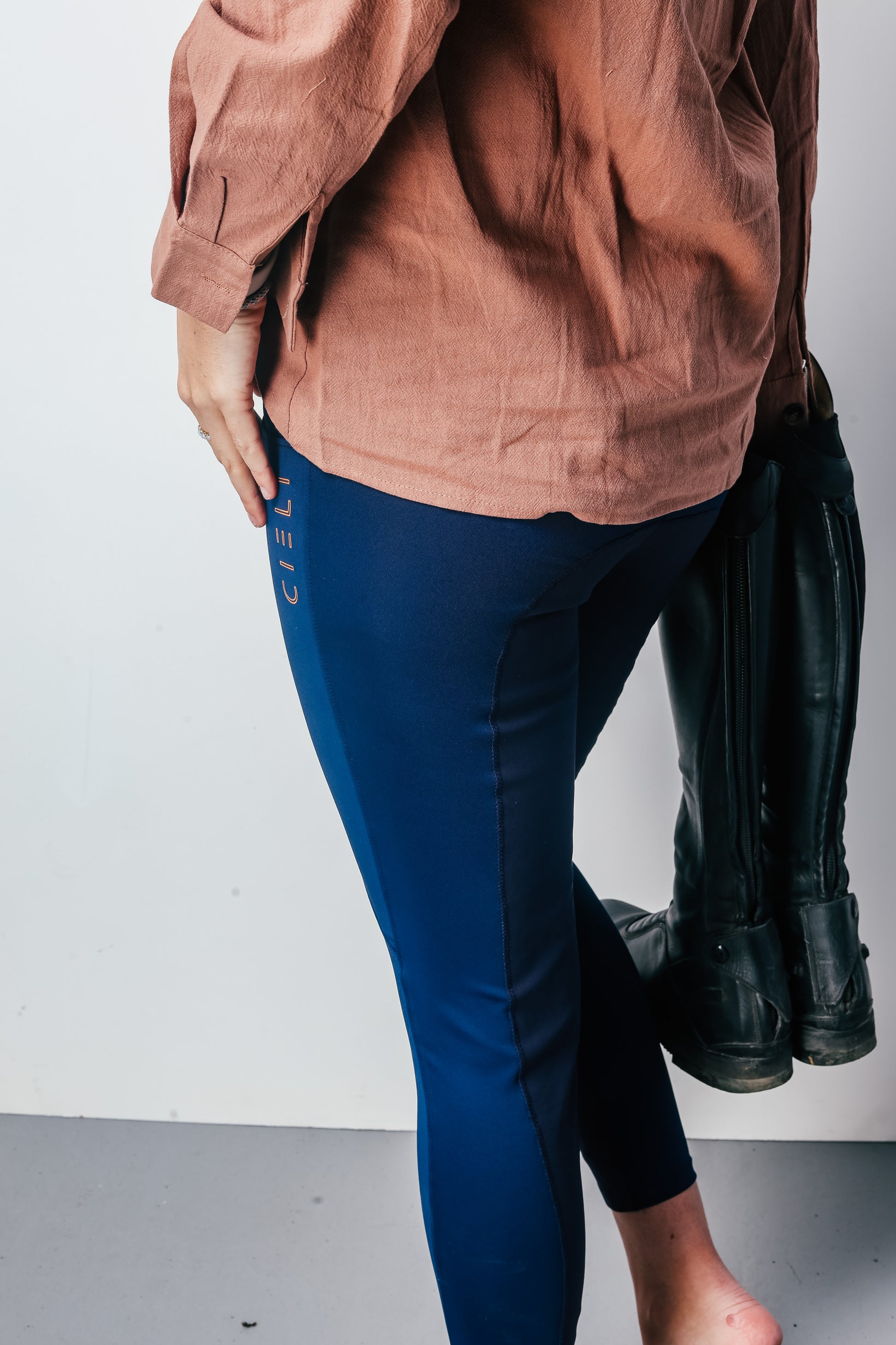 Person wearing a brown shirt and blue riding leggings with black boots on a white background