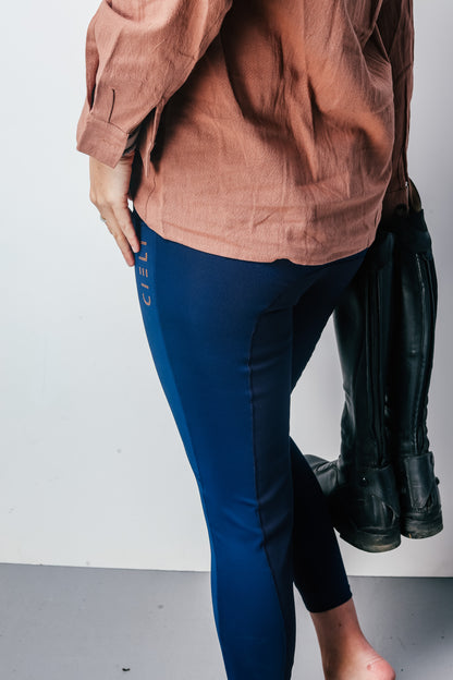 Person wearing a brown shirt and blue riding leggings with black boots on a white background