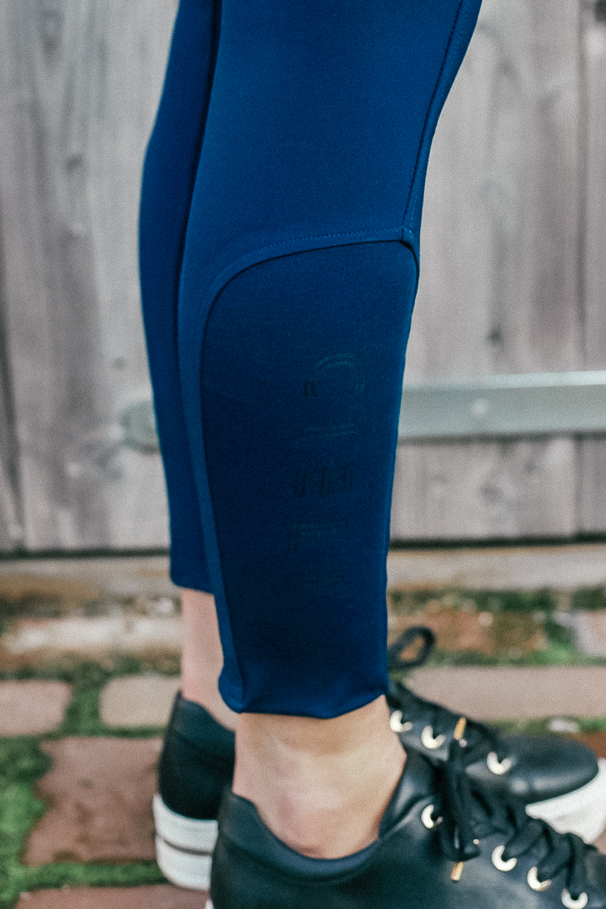 A close-up image showcases a person wearing the Chicago Everyday Breeches by Cieli Equestrian in bright blue, crafted from 4-way stretch fabric. They are paired with black sneakers, as the individual stands on a brick ground in front of a wooden gate. The breeches feature a small silicone print design near the calf area.