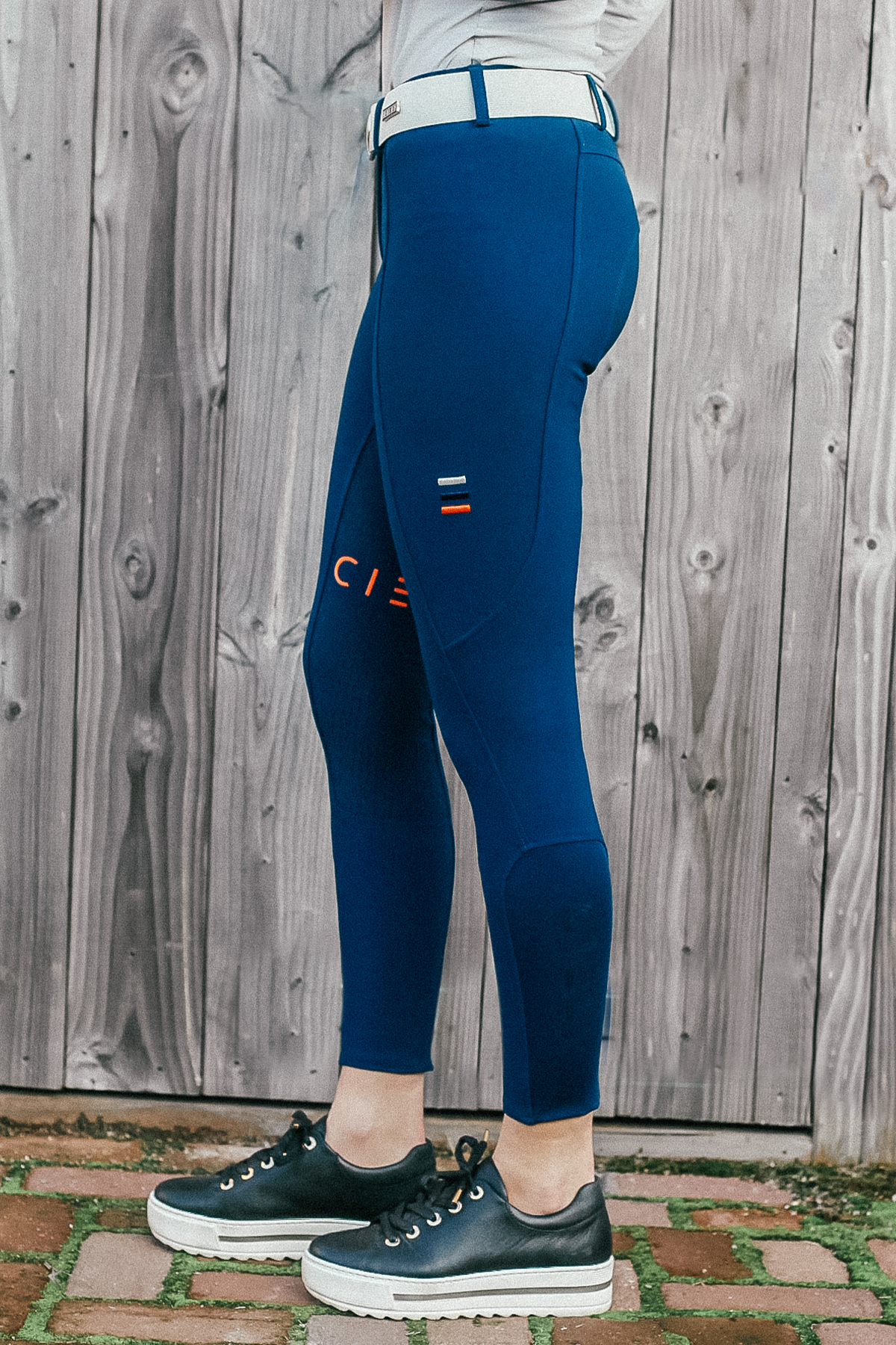 A person wearing vibrant blue Chicago Everyday Breeches from Cieli Equestrian and navy blue sneakers stands in profile against a wooden fence backdrop. Equestrian enthusiasts will appreciate the sporty red and white accents on the breeches' side, which feature 4-way stretch fabric for added comfort.