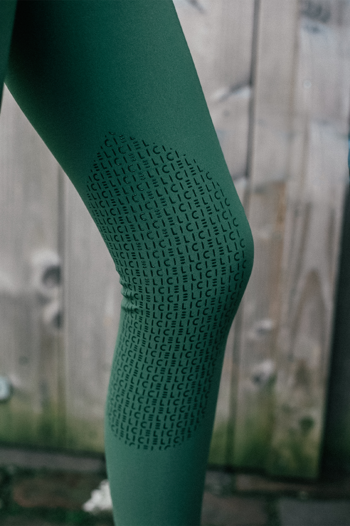 A close-up image of a person's knee wearing dark green leggings crafted from 4-way stretch fabric. These Chicago Everyday Breeches by Cieli Equestrian feature a prominent patch with an all-over "ICI" pattern in black. The background is softly blurred, revealing a wooden surface with some marks.