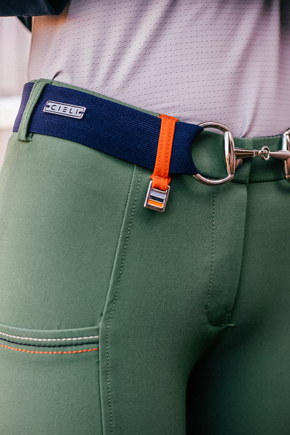 A close-up of a person wearing green Chicago Everyday Breeches by Cieli Equestrian with orange stitching and a navy blue belt that has a bright orange tab. The belt loop features a silver buckle and a small rectangular metal piece with the brand name "C4 BELT" engraved on it.