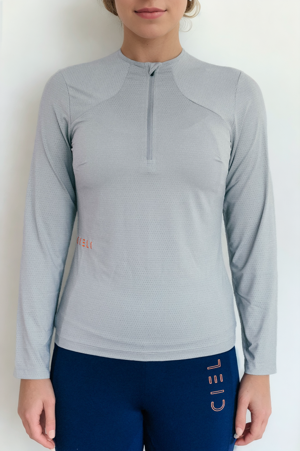 A person wears the Ella Baselayer from Cieli Equestrian, a light gray, lightweight, and breathable long-sleeved athletic top with a partial zip at the collar, paired with dark blue athletic pants. The background is plain white. The super soft fabric of the Ella Baselayer features subtle patterns and small orange text on the left chest area.