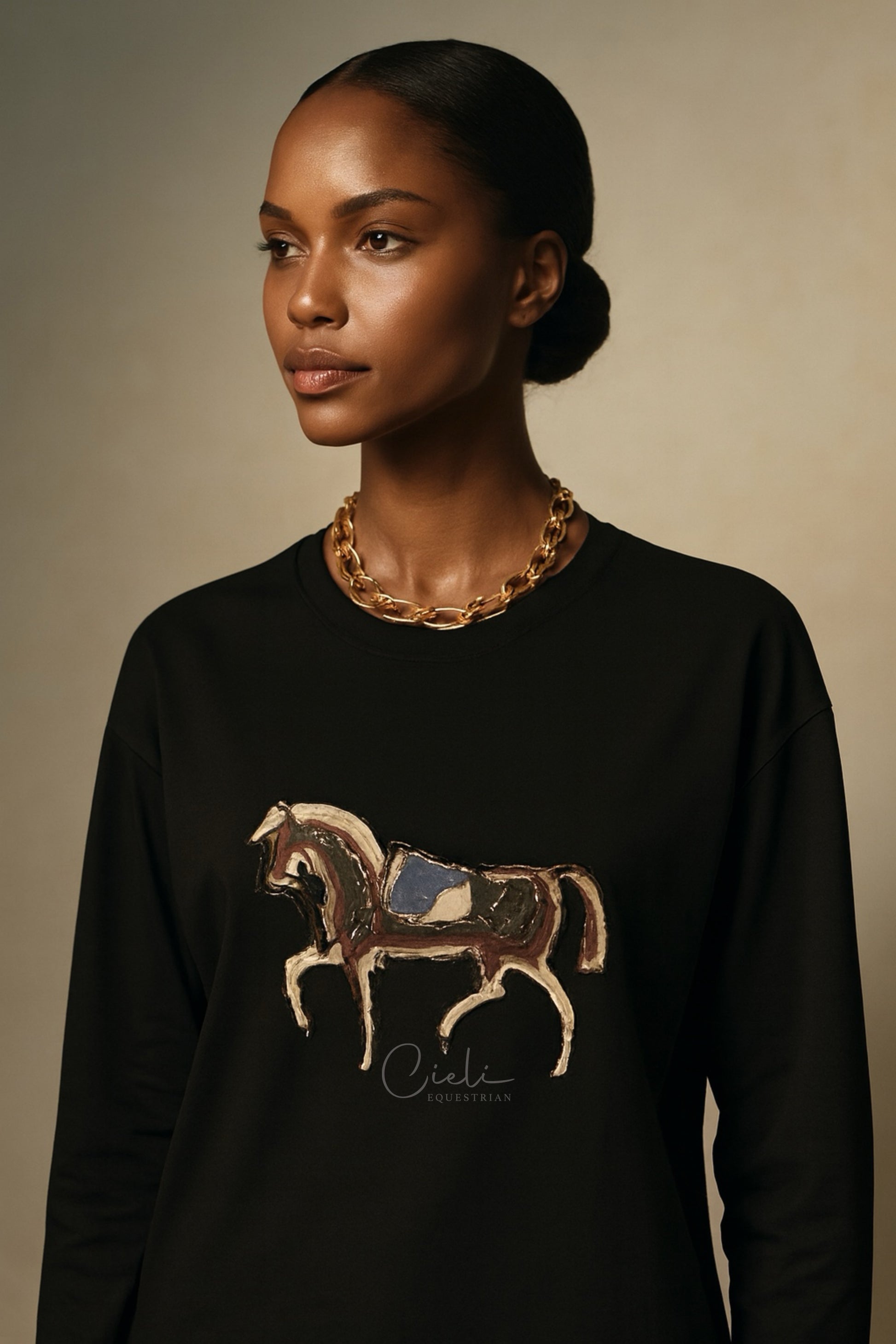 Woman wearing a black sweatshirt with a horse design and gold necklace against a beige background