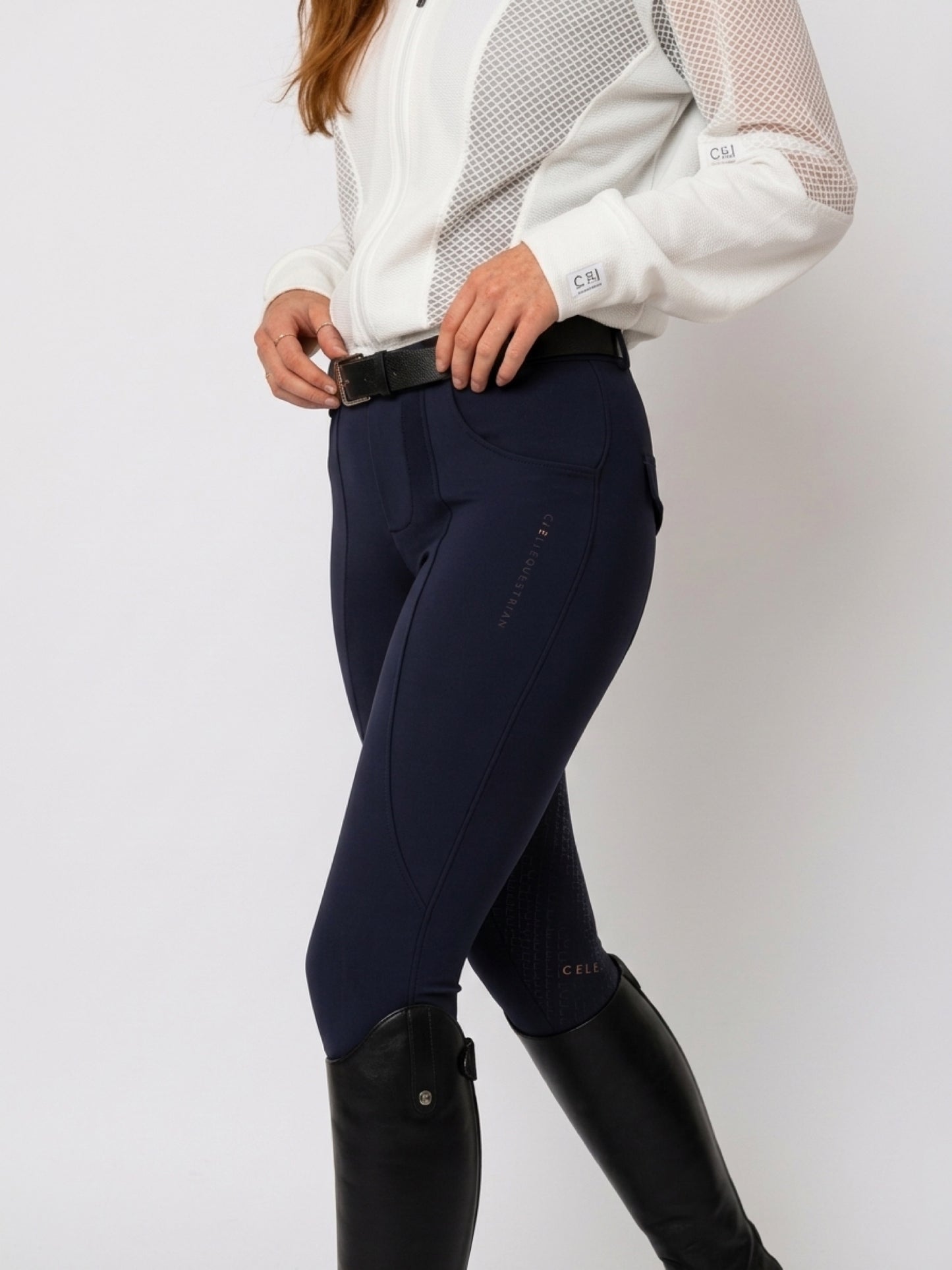 A model wearing navy blue equestrian riding breeches with a black belt, paired with a white long-sleeve equestrian shirt and black riding boots, shown from the waist down against a light gray studio background.