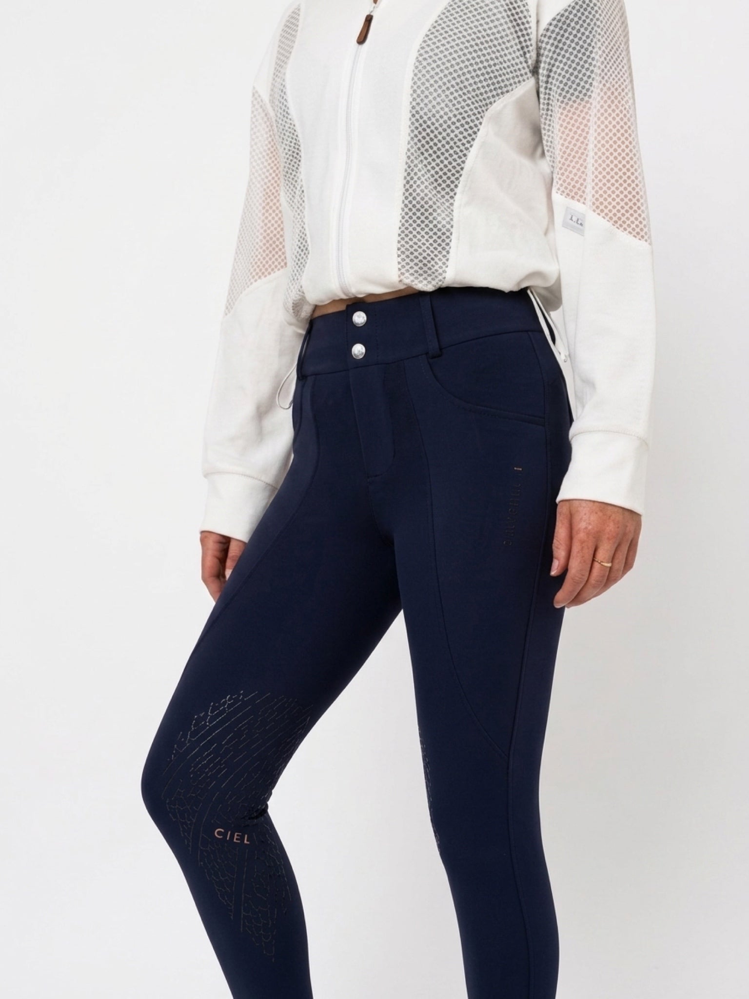 A model wearing navy blue equestrian riding breeches with textured knee panels and a small gold 'CIEL' logo near the lower left leg, paired with a white long-sleeve top with sheer mesh sleeves, against a plain white background.