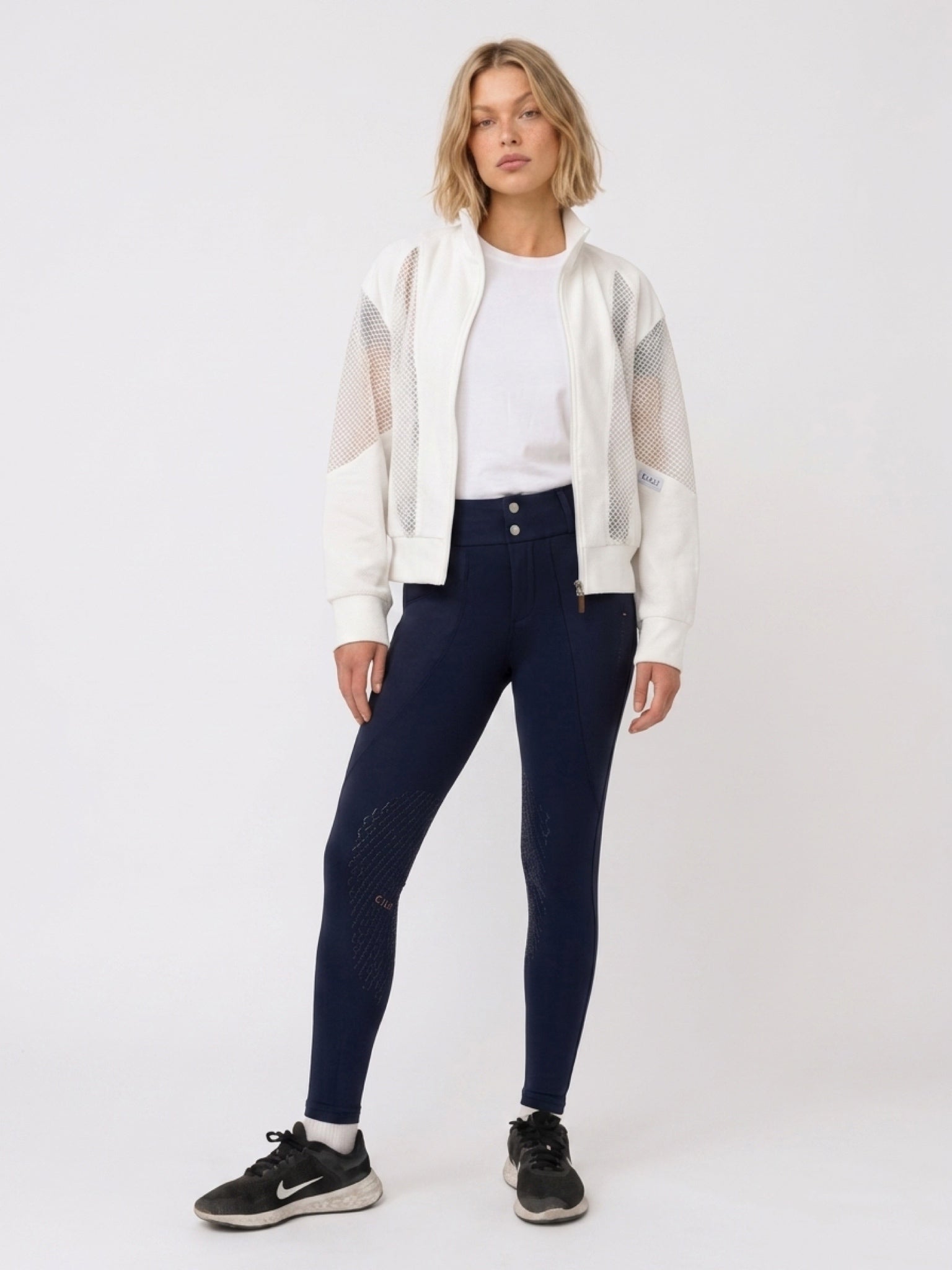 A woman modeling a pair of dark navy blue riding breeches, styled with a white t-shirt, a white cropped jacket with sheer sleeves, and black sneakers, against a plain white studio background.