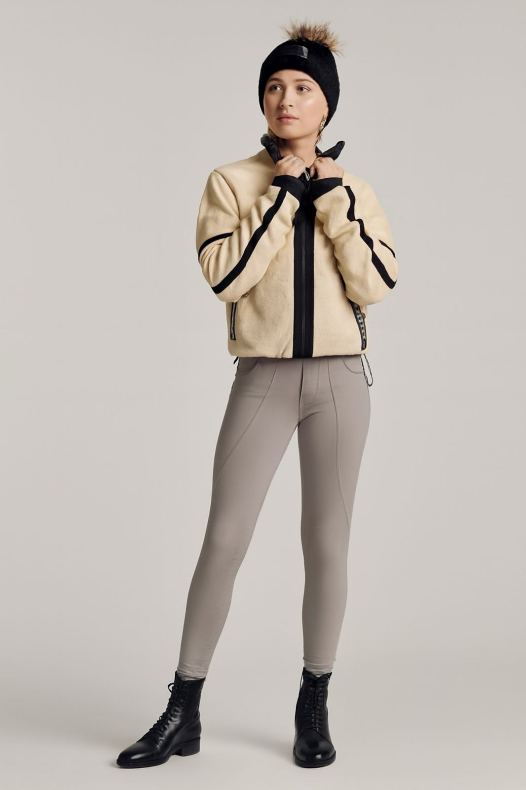 Person wearing a beige jacket with black accents, gray riding pants, and black boots on a plain background