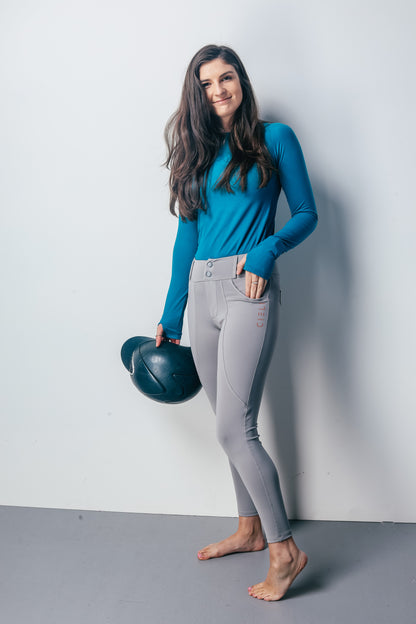 Woman wearing a blue top and gray riding leggings holding a helmet against a white background