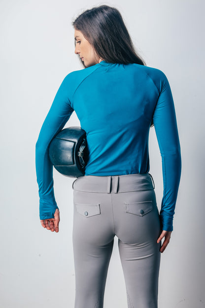 Woman wearing a blue top and gray riding pants holding a helmet against a white background