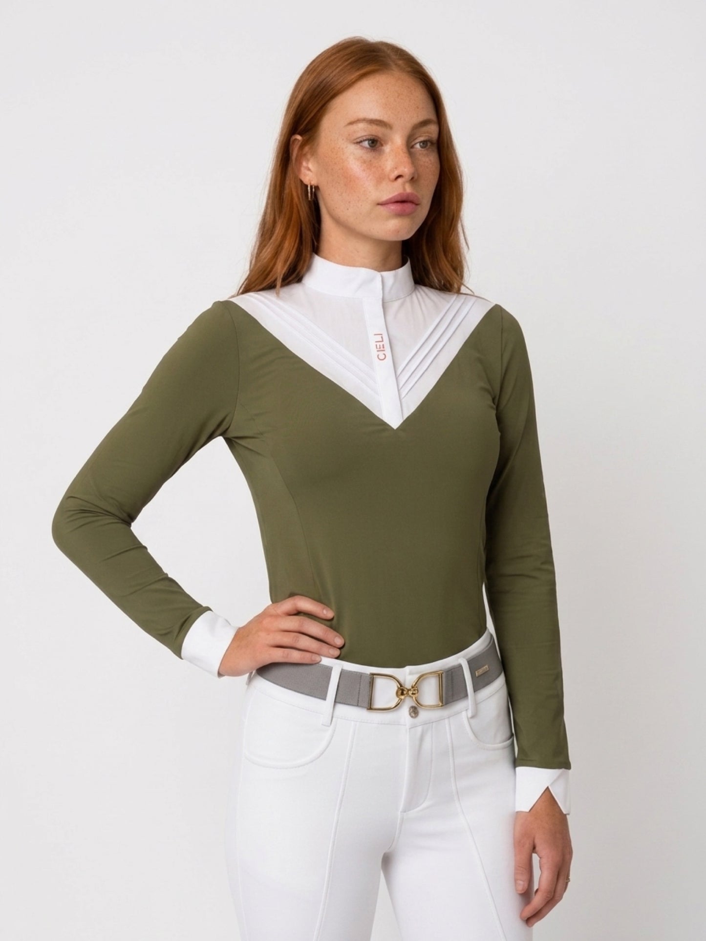 A woman modeling a fitted long-sleeve equestrian-style top in olive green with a contrasting white pleated V-neck collar and white cuffs, paired with white riding pants and a gray belt against a light gray studio background.