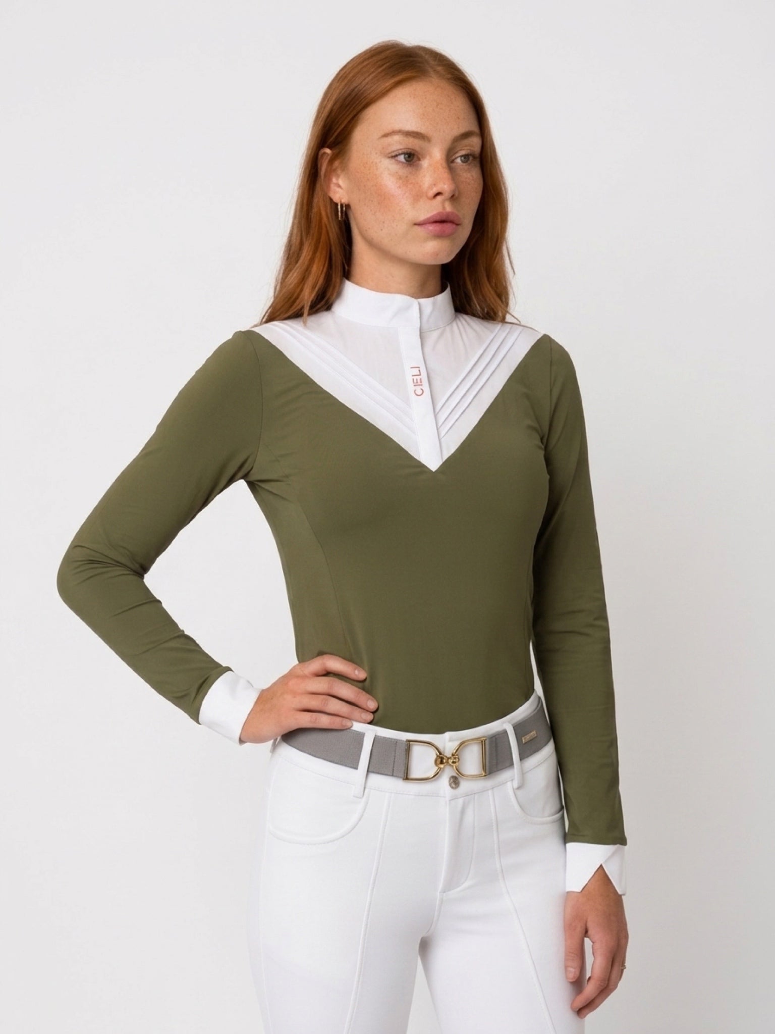 A woman modeling a fitted long-sleeve equestrian-style top in olive green with a contrasting white pleated V-neck collar and white cuffs, paired with white riding pants and a gray belt against a light gray studio background.