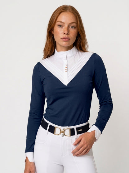 A woman modeling a navy blue long-sleeve show shirt with a contrasting white pleated V-neck collar, paired with white riding breeches and a black belt with a gold buckle, against a plain white studio background.