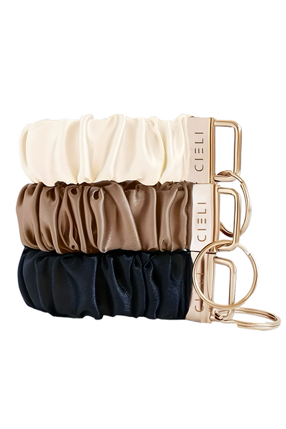 Set of three satin hair scrunchies in beige, brown, and navy with a gold holder on a white background.