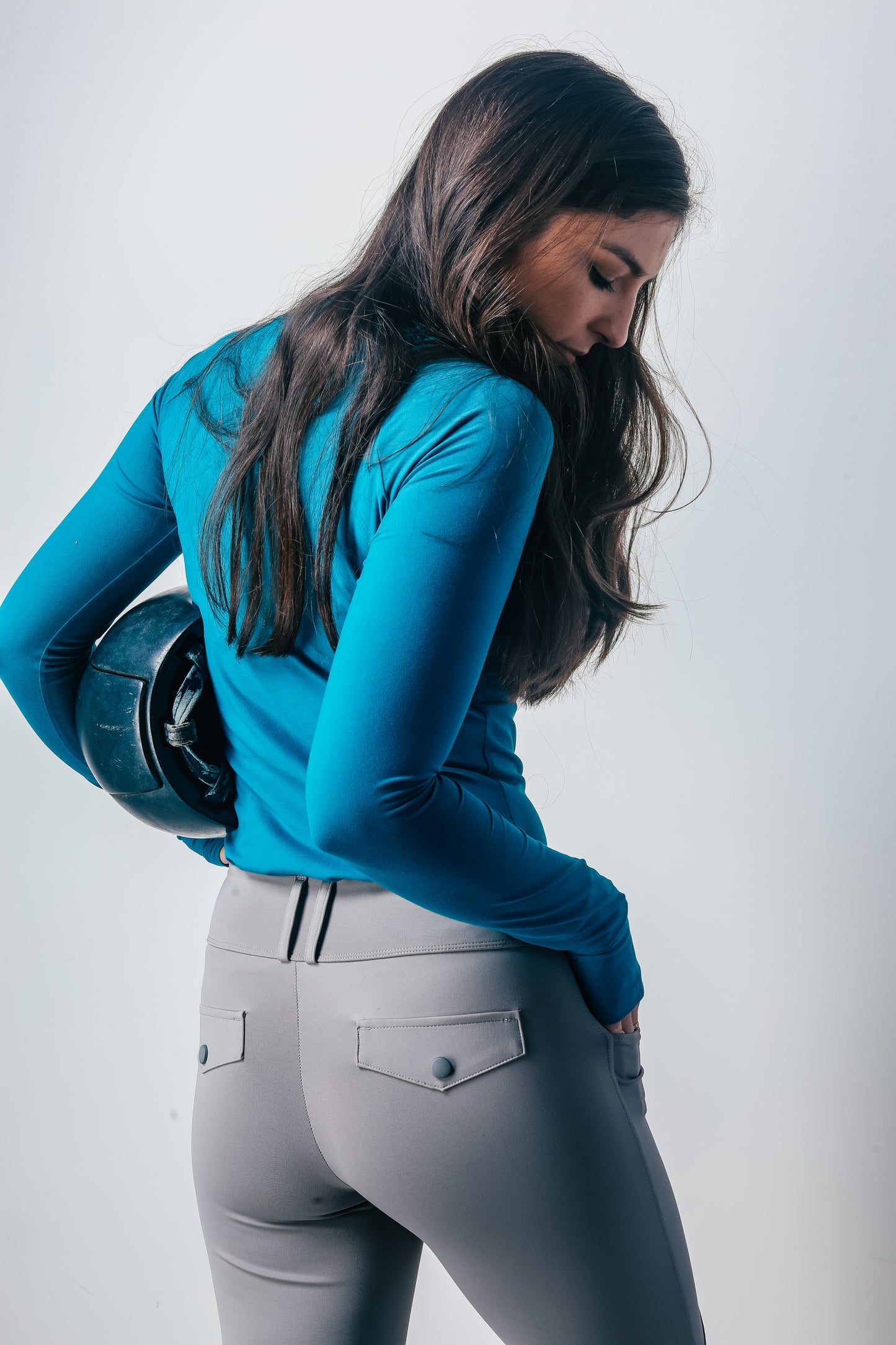 Woman wearing a blue top and gray riding pants holding a helmet against a white background