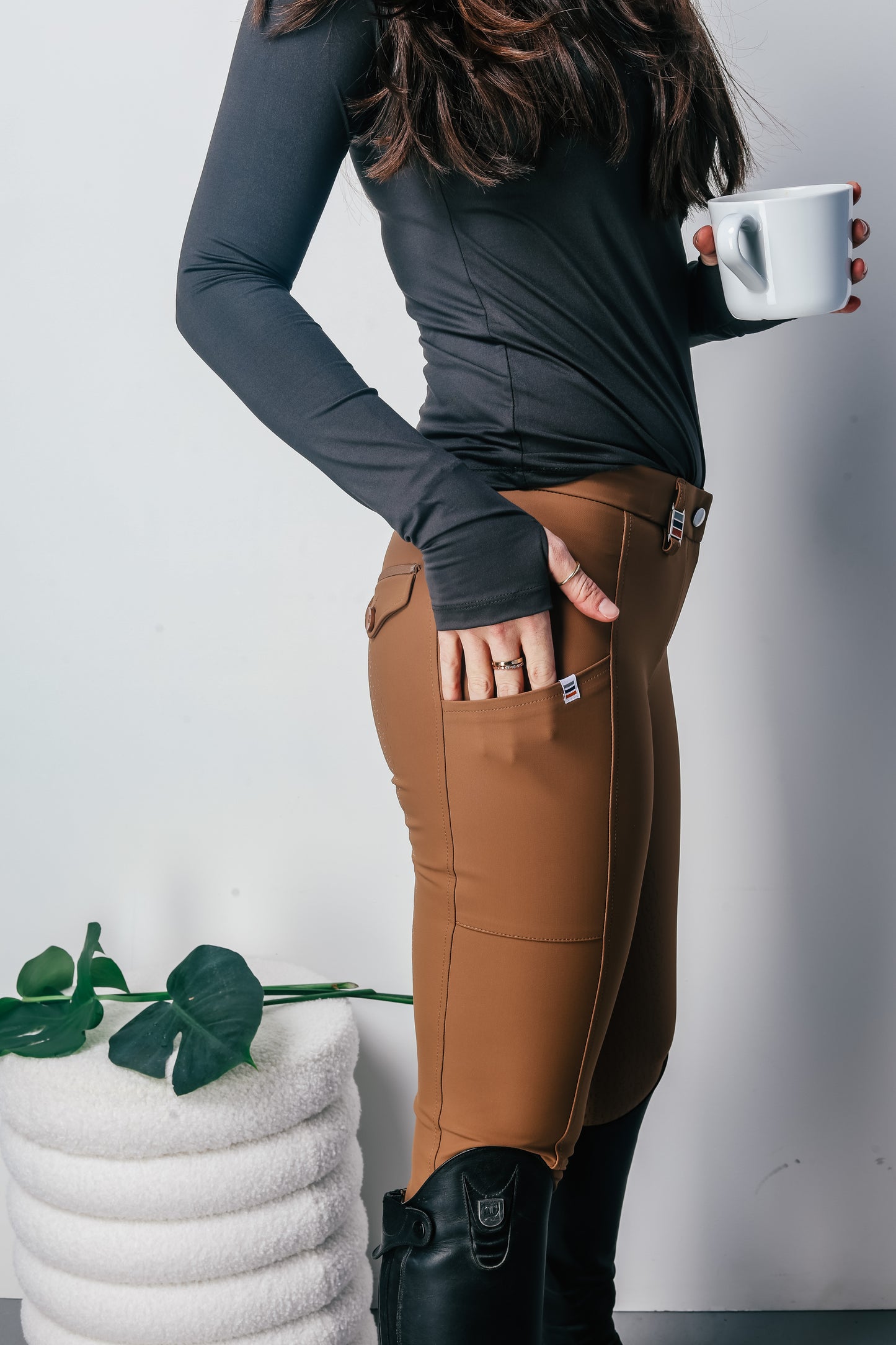 Person wearing brown riding pants and grey shirt holding a white mug with a plant in the background