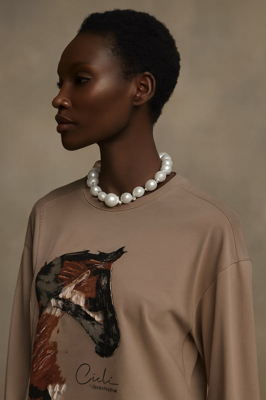 Woman wearing a beige sweatshirt with a horse design and pearl necklace against a neutral background