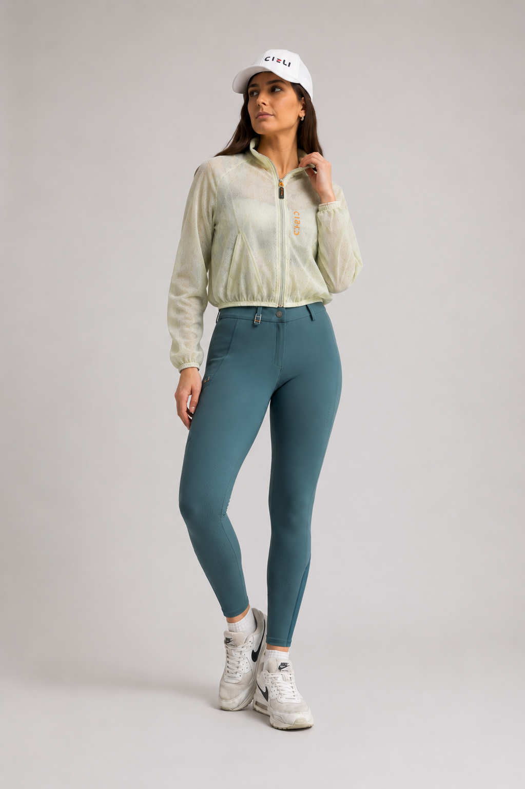 Woman wearing a light green jacket, teal riding breeches, and white sneakers on a plain background