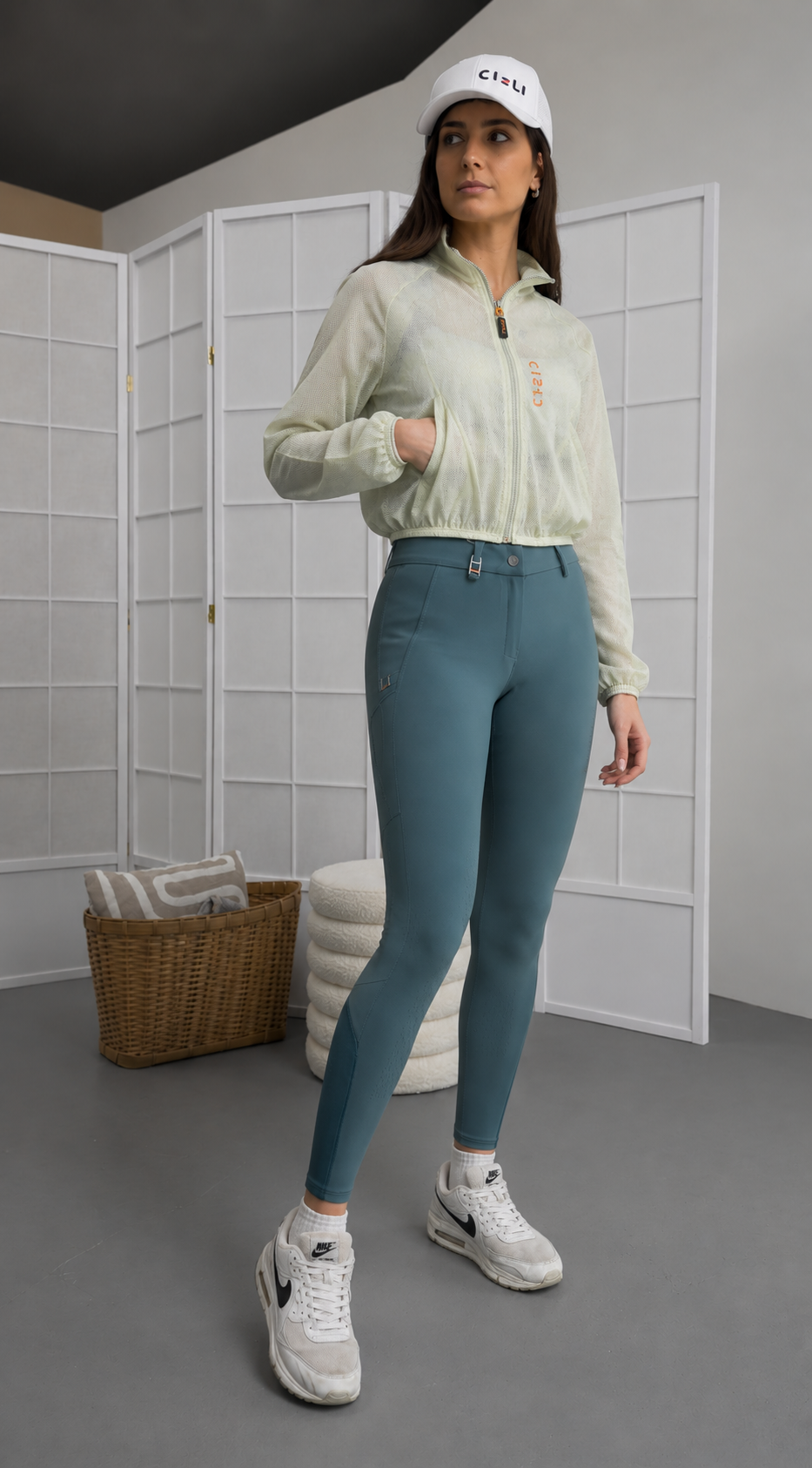 Woman wearing a light green jacket, blue riding breeches, and white sneakers in a room with a white wall and gray floor.