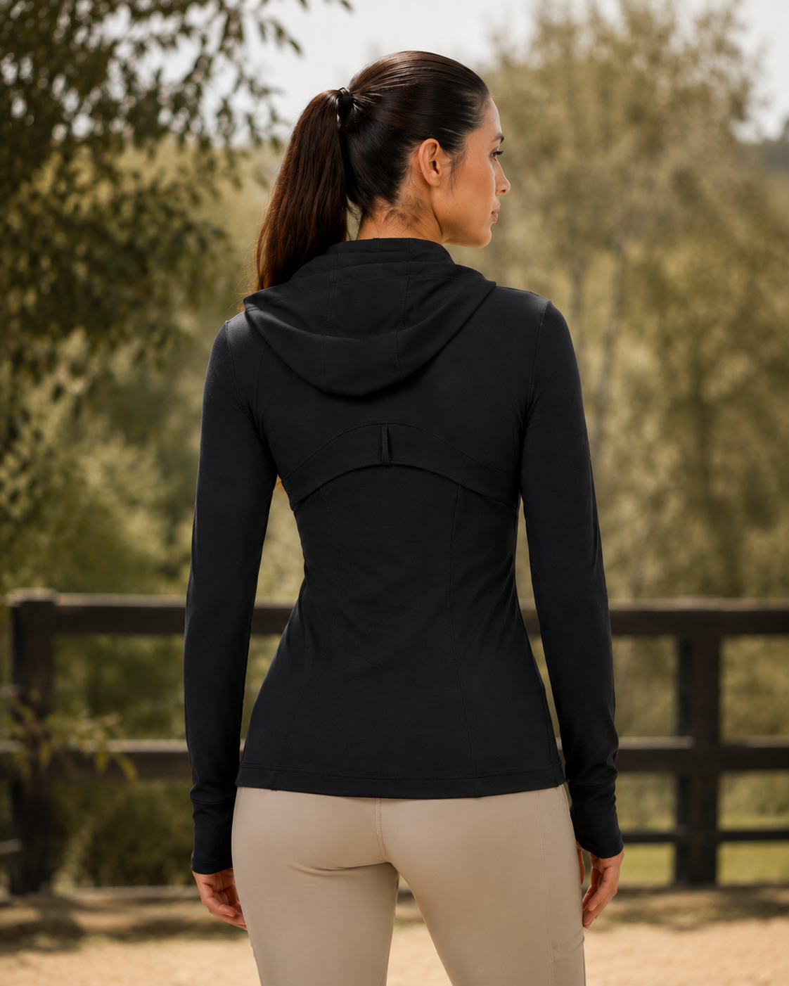A woman wearing a black long-sleeve athletic hooded top and beige leggings, shown from the back in an outdoor setting with trees and a fence in the background.